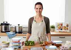 Un aperçu d'une cuisine moderne et lumineuse avec une femme souriante, devant un plan de travail où sont disposés des récipients de Batch Cooking colorés et organisés (légumes frais coupés, portions de plats préparés). L'ambiance est propre, efficace et inspirante.