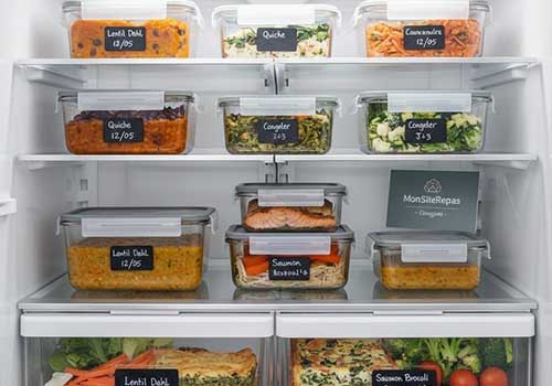 Un intérieur de réfrigérateur moderne et bien rangé. Sur les étagères, des boîtes de Batch Cooking transparentes et hermétiques, remplies de différents plats. Certaines boîtes ont de petites étiquettes avec la date de préparation ou le nom du plat, symbolisant les "Consignes de Conservation EXCLUSIVES". L'ensemble dégage une impression de propreté, d'ordre et de sécurité.