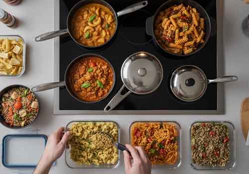 un plan de travail ou l"on peut voir différents plats dans des casseroles et les mains d'une personne en train de les repartir dans différents contenant hermétiques. 